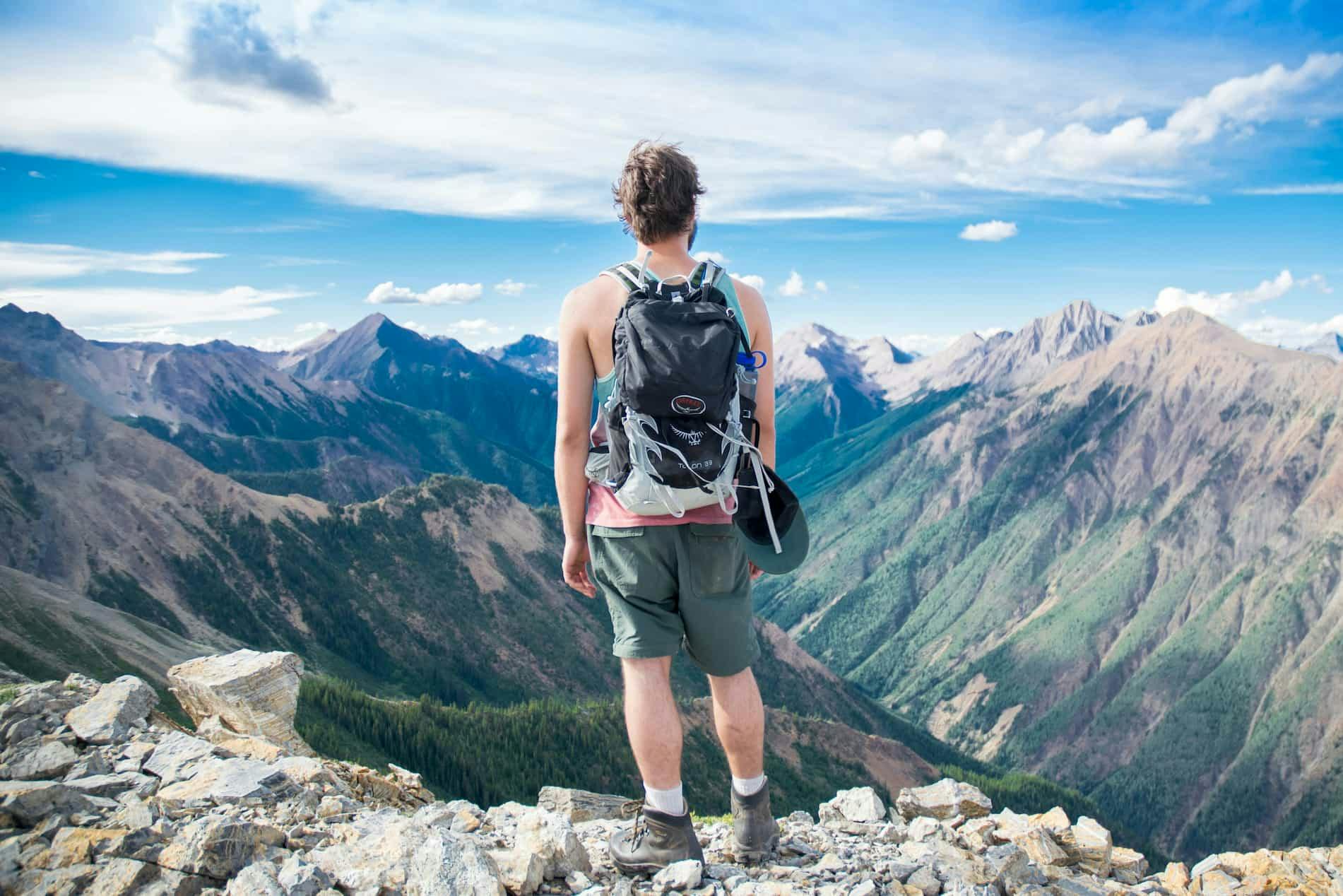 A backpacker stands on a rocky mountain peak, gazing at a vast landscape of rugged mountain ranges